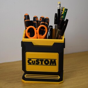 May include: A black and yellow desk organizer with the word "CUSTOM" on the front. The organizer holds various tools, including screwdrivers, scissors, pens, and pencils. The tools have black and orange handles.