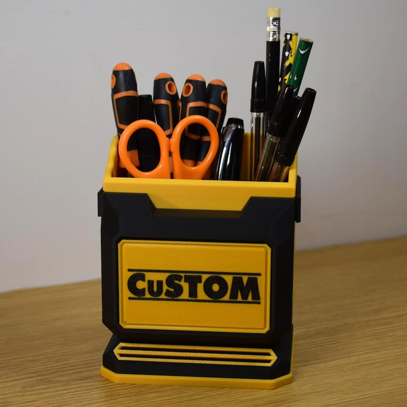 May include: A black and yellow desk organizer with the word "CUSTOM" on the front. The organizer holds various tools, including screwdrivers, scissors, pens, and pencils. The tools have black and orange handles.