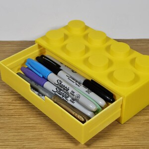 May include: A yellow plastic drawer organizer shaped like a Lego brick. The drawer is open and contains a variety of colored markers, including black, blue, purple, and green.