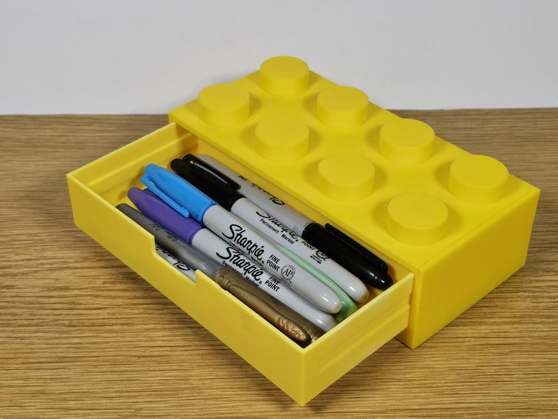 May include: A yellow plastic drawer organizer shaped like a Lego brick. The drawer is open and contains a variety of colored markers, including black, blue, purple, and green.