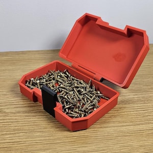 Op de afbeelding: Een rode plastic opbergdoos met een zwart slot, open om een ​​grote hoeveelheid gouden schroeven te laten zien.