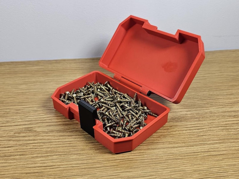Op de afbeelding: Een rode plastic opbergdoos met een zwart slot, open om een ​​grote hoeveelheid gouden schroeven te laten zien.