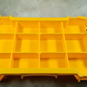 May include: A bright yellow storage container with multiple compartments. The container has a rectangular shape with a series of dividers creating individual storage spaces. Black handles are on each side. Ideal for organizing small items.