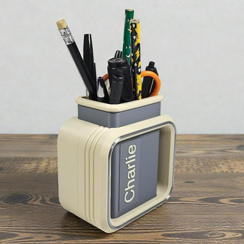 May include: A cream and gray desk organizer with the name "Charlie" printed on the front. The organizer holds various pens, pencils, and scissors. The pencils have yellow, green, and black designs.