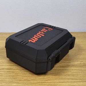 May include: Black rectangular box with the word "Custom" in orange lettering on the lid. The box has a hinged lid and a latch. The box is sitting on a wooden surface.