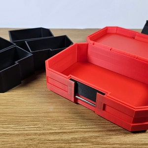 May include: A red plastic storage box with a black latch and a black interior. The box is open and empty. There are three black plastic storage containers in the background.