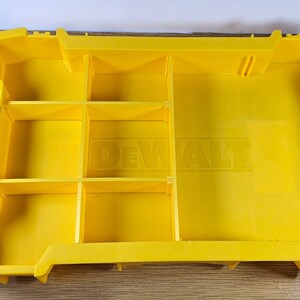 Op de afbeelding: Gele plastic gereedschapskist met meerdere vakken en het woord "DEWALT" in reli&euml;f op de bodem. De kist is aan de zijkant ge&euml;tiketteerd met "TOUGHSYSTEM".