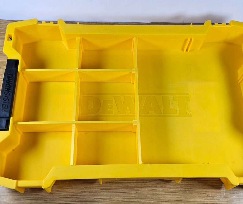 Op de afbeelding: Gele plastic gereedschapskist met meerdere vakken en het woord "DEWALT" in reli&euml;f op de bodem. De kist is aan de zijkant ge&euml;tiketteerd met "TOUGHSYSTEM".
