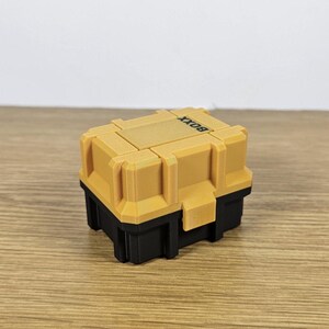 May include: A small, yellow and black box with the word "BOXX" printed on the top. The box has a textured surface and appears to be made of plastic.