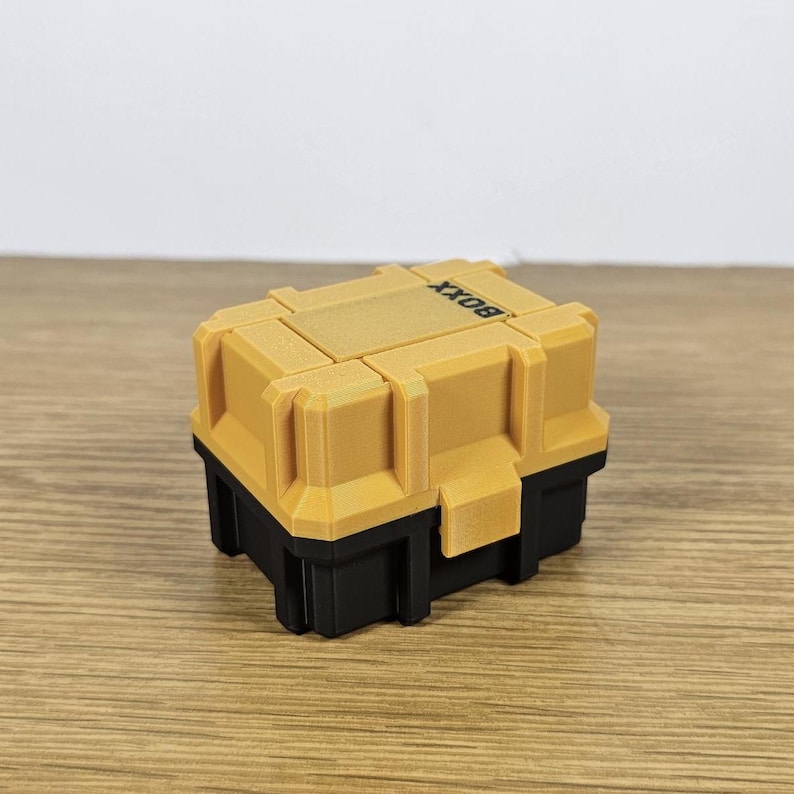 May include: A small, yellow and black box with the word "BOXX" printed on the top. The box has a textured surface and appears to be made of plastic.