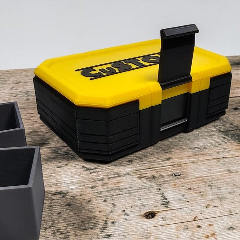 May include: A yellow and black toolbox with the word "CUSTOM" printed on the lid. The toolbox has a handle and is sitting on a wooden surface. The toolbox is rectangular and has a black base.