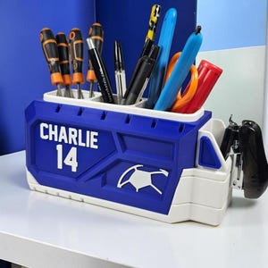 May include: A blue and white plastic desk organizer shaped like a truck, featuring the name CHARLIE and the number 14 in white text. The organizer holds various office supplies, including screwdrivers, pens, and a black stapler attached to the side.
