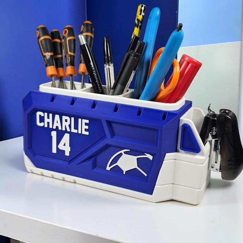 May include: A blue and white plastic desk organizer shaped like a truck, featuring the name CHARLIE and the number 14 in white text. The organizer holds various office supplies, including screwdrivers, pens, and a black stapler attached to the side.