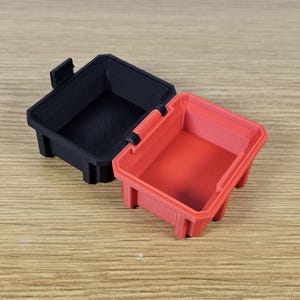 May include: Two small, rectangular storage containers. One is black, the other is red. Both have a handle and a latch. The containers are made of a solid material and are designed to hold small items. The containers are sitting on a wooden surface.