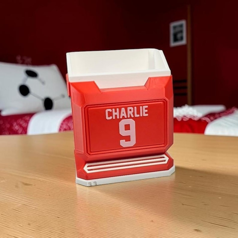 May include: A red and white desk organizer shaped like a sports jersey, featuring the name CHARLIE and the number 9 printed in white on the front. The container has a rectangular opening at the top and a structured, angular design.