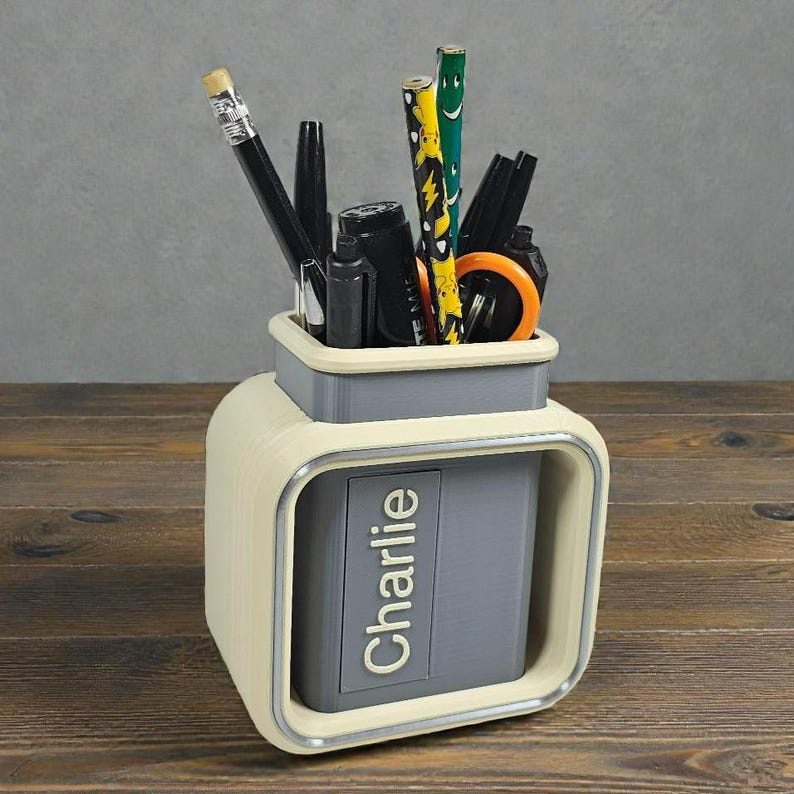 May include: A cream and gray desk organizer with the name "Charlie" printed on the front. The organizer holds pencils, pens, and scissors. The desk organizer is on a wooden surface.