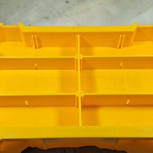 May include: A bright yellow plastic tool organiser with multiple compartments. The organiser has a rectangular shape with several dividers creating storage sections. It features a handle and a secure closure system for easy transport and organisation.