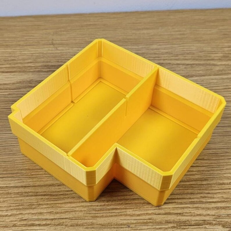 May include: A bright yellow, corner-shaped organizer with two compartments. The organizer is made of a solid material and has a light yellow trim around the top edges. The design is simple and functional, suitable for desk or drawer organization.