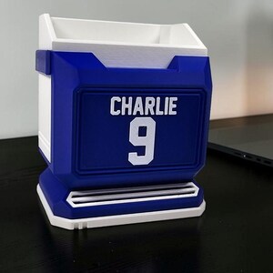 May include: A blue and white desk organizer with the name "CHARLIE" and the number "9" in white. The organizer has a rectangular shape with a slot at the bottom and a compartment at the top.
