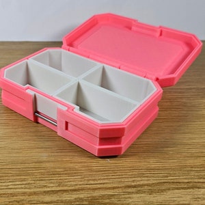 May include: A pink plastic pill box with four compartments. The box is open and the compartments are empty.
