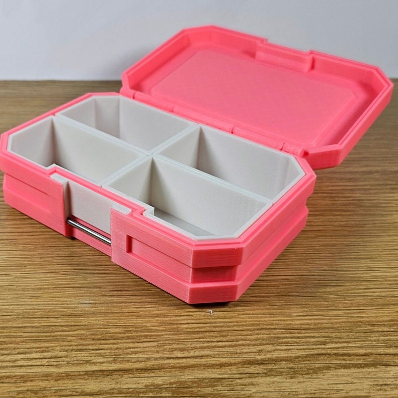May include: A pink plastic pill box with four compartments. The box is open and the compartments are empty.