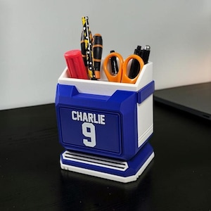 May include: A blue and white desk organizer with the name "Charlie" and the number "9" on the front. The organizer holds pens, pencils, scissors, and a red marker. The design is geometric and modern.
