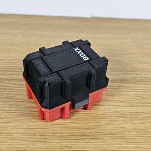 Pu&ograve; includere: Una piccola scatola di plastica nera e rossa con la scritta "BOX" stampata sulla parte superiore.