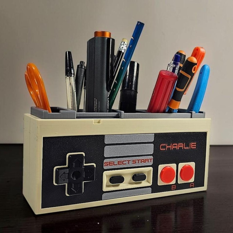 May include: A beige and black desk organizer shaped like a classic video game controller, holding pens, scissors, and other office supplies. The text "CHARLIE" is printed on the front. The controller design includes a directional pad, buttons, and the words "SELECT START".