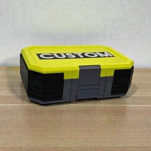 May include: A rectangular storage box with a bright yellow lid and black body. The lid features the word "CUSTOM" in bold white letters. The box has gray accents and is sitting on a wooden surface.