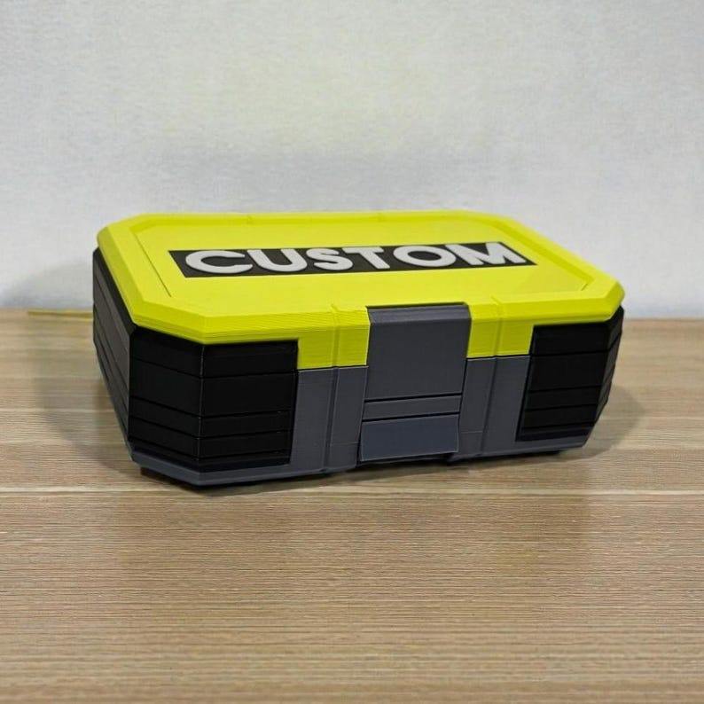 May include: A rectangular storage box with a bright yellow lid and black body. The lid features the word "CUSTOM" in bold white letters. The box has gray accents and is sitting on a wooden surface.
