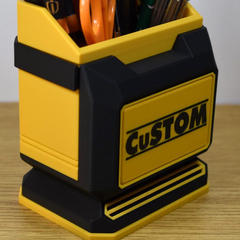 May include: A yellow and black desk organizer with the word "CUSTOM" in white. The organizer is filled with pens and orange scissors. The design features a rectangular shape with a base and a handle-like detail.