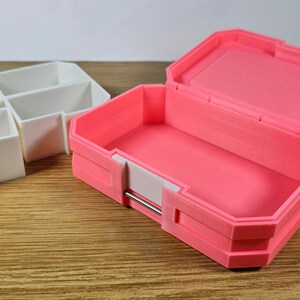 May include: Two small storage boxes, one white and one pink. The white box has four compartments. The pink box is open and has a white latch.