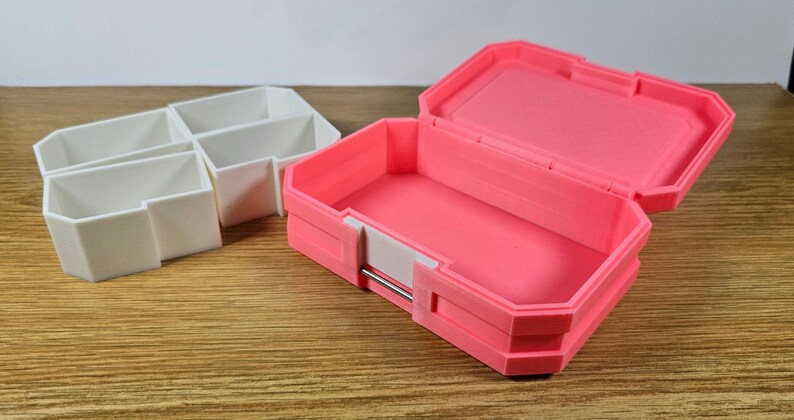 May include: Two small storage boxes, one white and one pink. The white box has four compartments. The pink box is open and has a white latch.