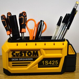 K&ouml;nnte beinhalten: Ein gelb-schwarzer Schreibtisch-Organizer mit dem Text "CUSTOM DESK ORGANISER" und "1S425". Der Organizer enth&auml;lt Schraubendreher, Scheren, Stifte und Bleistifte. Die Werkzeuge haben orange und schwarze Akzente.