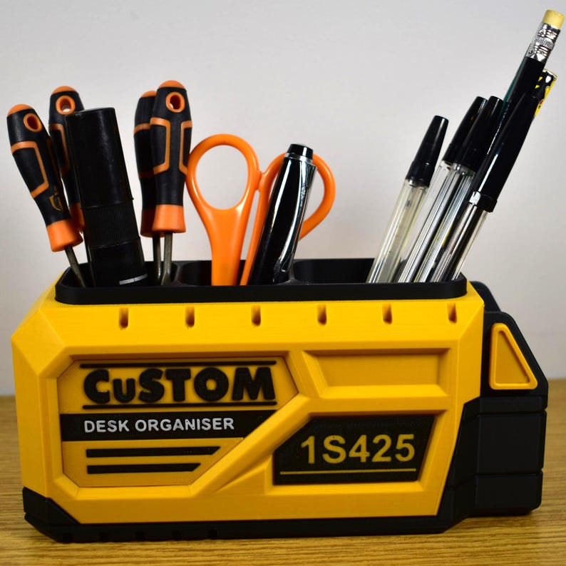 K&ouml;nnte beinhalten: Ein gelb-schwarzer Schreibtisch-Organizer mit dem Text "CUSTOM DESK ORGANISER" und "1S425". Der Organizer enth&auml;lt Schraubendreher, Scheren, Stifte und Bleistifte. Die Werkzeuge haben orange und schwarze Akzente.