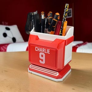 May include: A red and white desk organizer shaped like a sports jersey, featuring the name CHARLIE and the number 9 printed on the front. The container holds various office supplies, including pens, a stapler, and a pencil with a yellow pattern.
