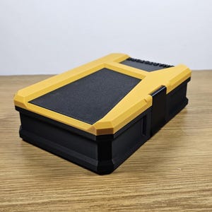 May include: A small, black and yellow storage box with a textured lid. The box has a latch on the side.