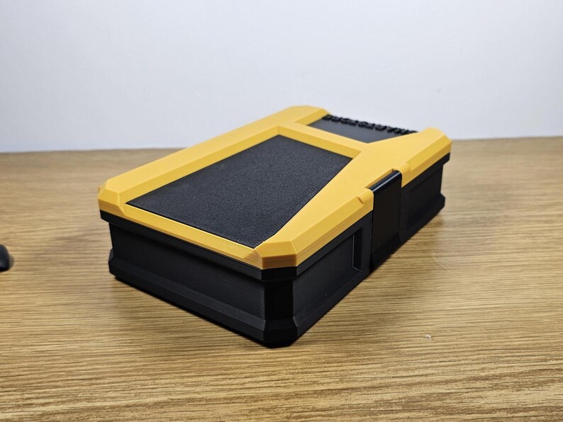 May include: A small, black and yellow storage box with a textured lid. The box has a latch on the side.