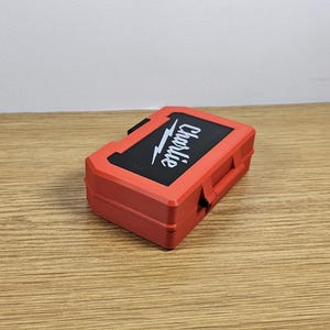 Op de afbeelding: Een rode plastic gereedschapskist met een zwart deksel en het woord "Charlie" in wit op het deksel.