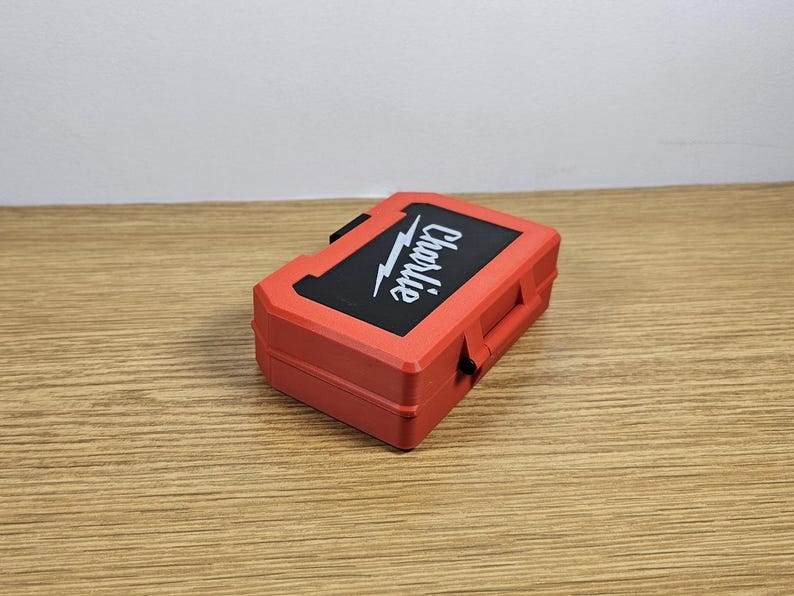 Op de afbeelding: Een rode plastic gereedschapskist met een zwart deksel en het woord "Charlie" in wit op het deksel.