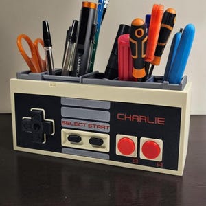 May include: A desk tidy in the shape of a retro games console controller, in beige and black. It holds pens, scissors, and screwdrivers. The name "CHARLIE" is printed in red, and the controller buttons are visible.