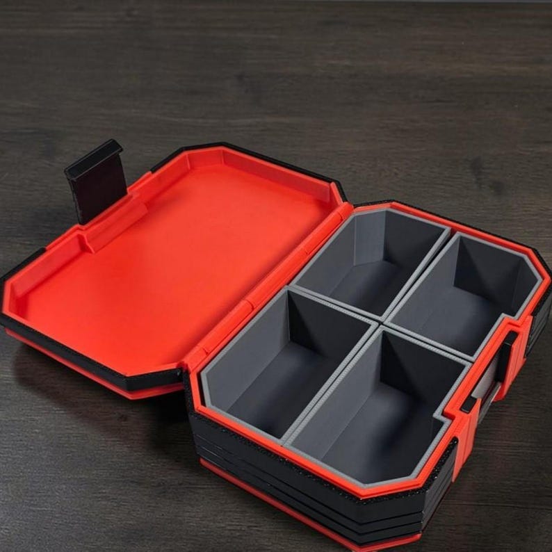 May include: Open storage box with multiple compartments. The box has a red interior and a black and grey exterior. It is rectangular with a hinged lid and a latch. The interior is divided into four grey compartments.