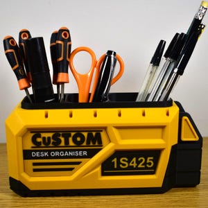 K&ouml;nnte beinhalten: Ein gelb-schwarzer Schreibtisch-Organizer mit den W&ouml;rtern "CUSTOM" und "DESK ORGANISER" auf der Vorderseite. Er enth&auml;lt Schraubendreher, Scheren, Stifte und Bleistifte. Die Modellnummer "1S425" ist ebenfalls aufgedruckt.