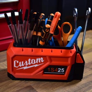 May include: A red and black tool organizer with the word "Custom" in white. The organizer holds pens, screwdrivers, scissors, and wrenches. The text "1S425" is also visible on the organizer.