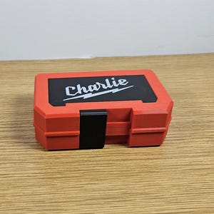 Op de afbeelding: Rode plastic gereedschapskist met een zwart slot en het woord "Charlie" in wit op de bovenkant.
