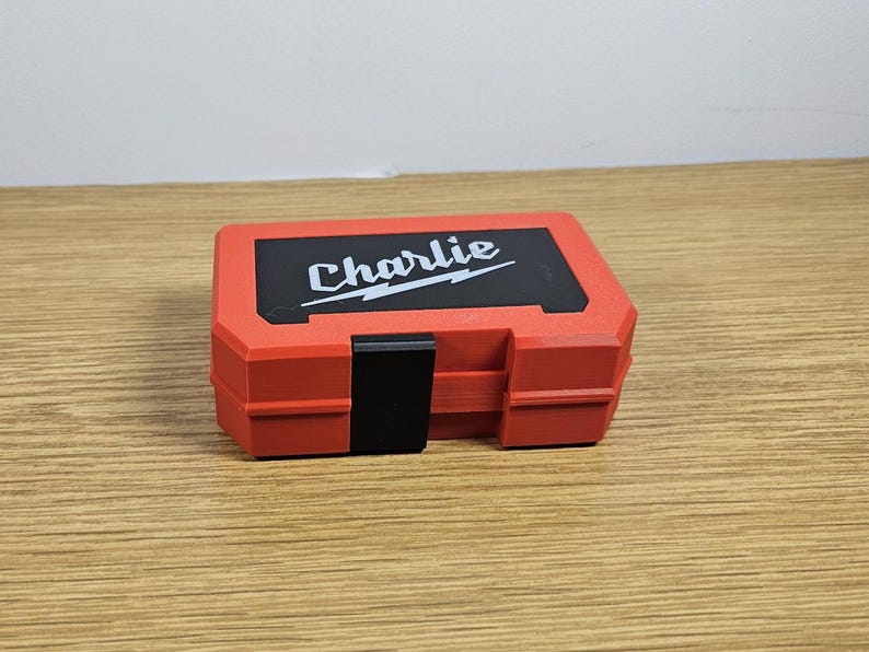 Op de afbeelding: Rode plastic gereedschapskist met een zwart slot en het woord "Charlie" in wit op de bovenkant.