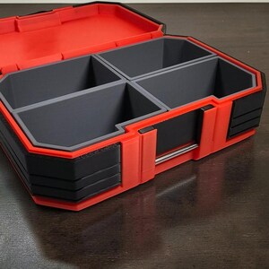 May include: A red and black storage container with four grey compartments. The container is open, revealing the interior layout. The exterior features a combination of red and black plastic, with a silver metal handle. The design is angular and modern.