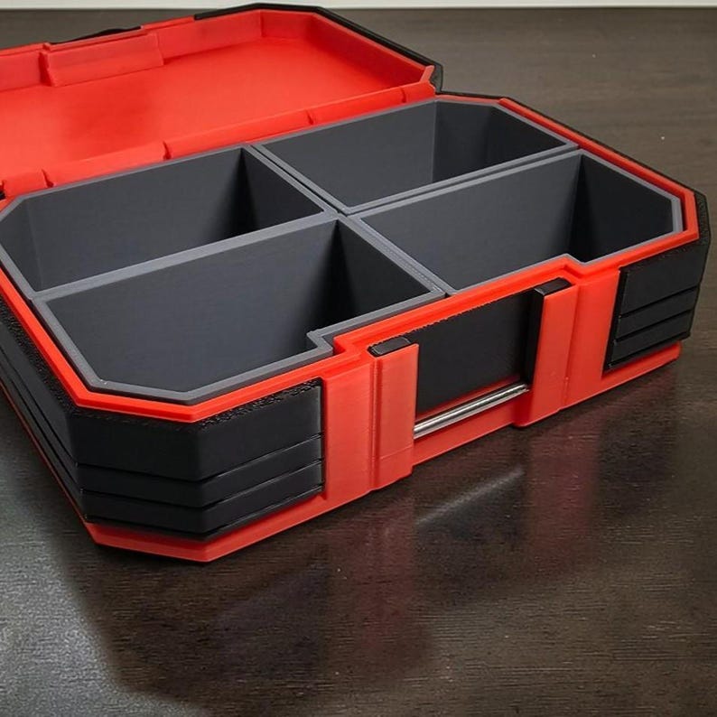 May include: A red and black storage container with four grey compartments. The container is open, revealing the interior layout. The exterior features a combination of red and black plastic, with a silver metal handle. The design is angular and modern.