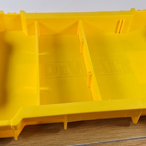 Op de afbeelding: Gele plastic opbergdoos met verdelers en een zwarte handgreep. De doos heeft het woord "DEWALT" op de bodem.
