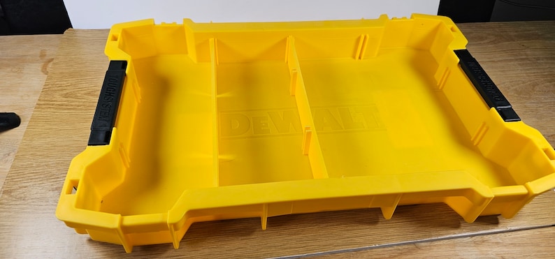 Op de afbeelding: Gele plastic opbergdoos met verdelers en een zwarte handgreep. De doos heeft het woord "DEWALT" op de bodem.